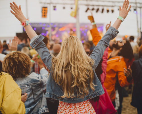 Young woman with blonde hair is dancing with her friends in a performance tent at a music festival. She has her arms outstretched and is watching the performance.